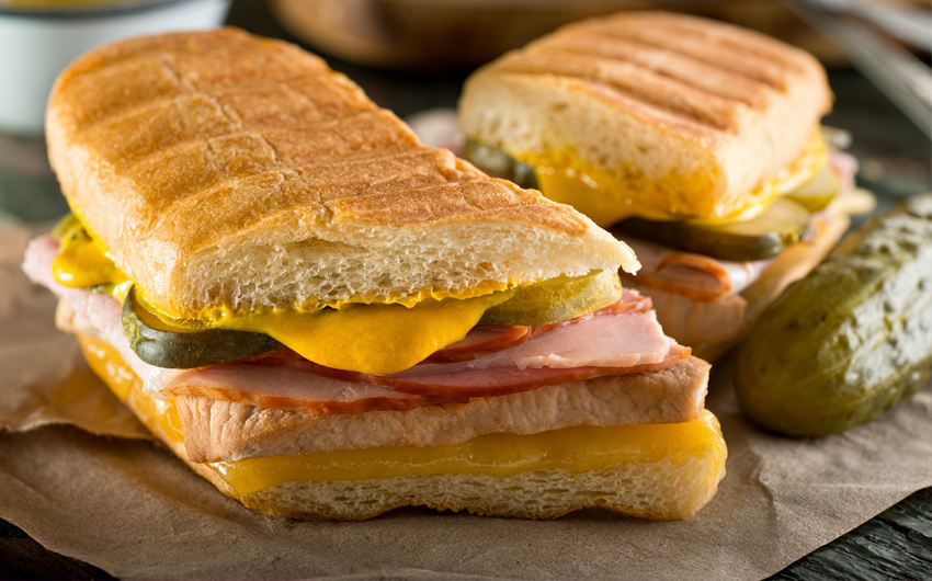 An authentic cuban sandwich on pressed medianoche bread with pork, ham, cheese, pickle, and mustard